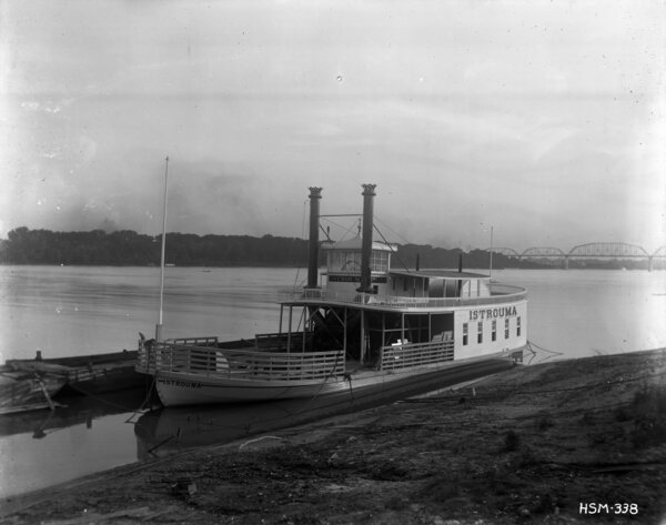 Photograph taken from the bank showing the Istrouma ferry boat docked along the bank of the Mississippi River with a bridge in the background crossing the river.